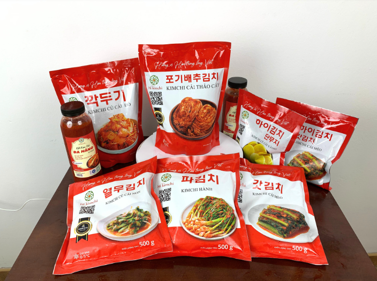 Ảnh chụp sản phẩm Hi Kim Chi của Pusan Foods. Hình ảnh cần thể hiện hũ/bao bì sản phẩm rõ ràng, sạch sẽ, có nhãn mác chuyên nghiệp. Đặt sản phẩm trong bối cảnh bếp ăn gia đình hiện đại hoặc bên cạnh các món ăn Việt Nam (ví dụ: bên cạnh bát mì gói, hoặc đĩa thịt luộc) để thể hiện sự tiện lợi và phù hợp. Nên có logo Pusan Foods ở góc.