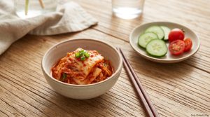 Một bát kim chi cải thảo đỏ rực rỡ đặt cạnh một quả cà chua hoặc vài lát dưa chuột trên mặt bàn gỗ. Có thể thêm một dòng text nhỏ, tinh tế trên ảnh: "Chỉ 20 kcal/100g".