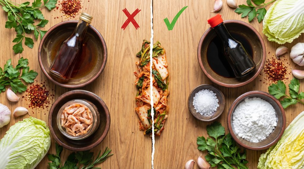 Ảnh chụp flatlay (từ trên xuống) chia làm 2 bên. Một bên là các nguyên liệu truyền thống (có mắm cá, tép muối - gạch chéo đỏ nhẹ). Một bên là nguyên liệu chay (nước tương, muối biển, bột gạo - dấu tích xanh). Hình ảnh này giúp người đọc nhận diện ngay "insight" tại sao kim chi thường không chay.