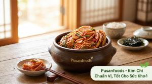 Một mẹt kim chi cải thảo đỏ tươi, trang trí đẹp mắt với mè trắng. Có logo Pusanfoods ở góc.
