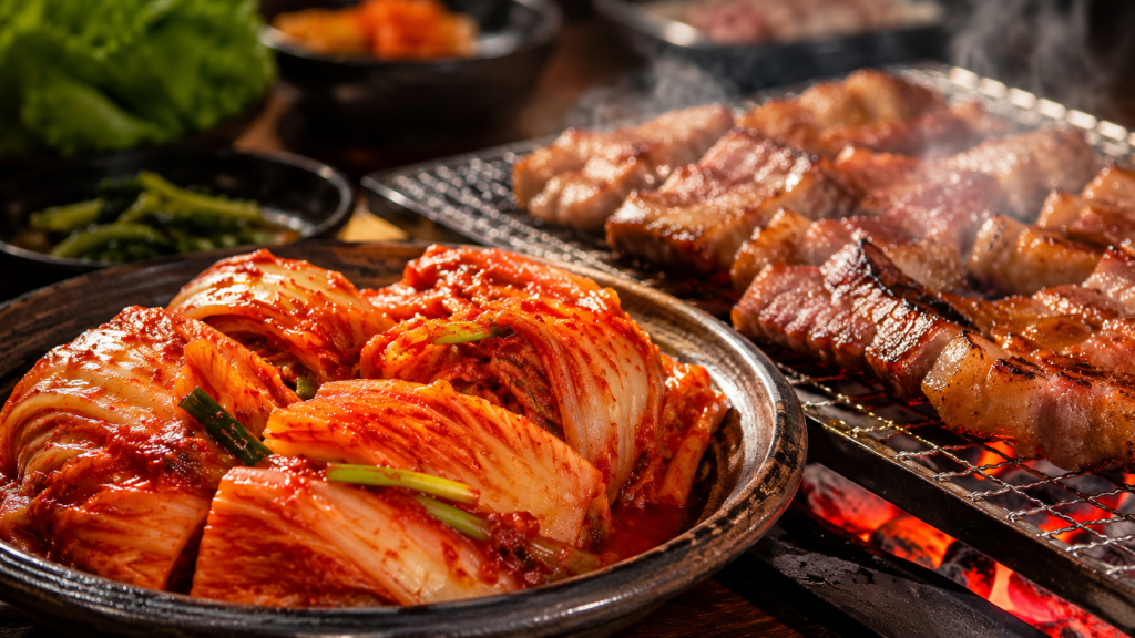 Một đĩa kim chi cải thảo cắt khúc, màu đỏ mọng, đặt cạnh một đĩa thịt ba chỉ nướng cháy cạnh trên bếp than hồng. Khói bốc lên nhẹ, tạo cảm giác cực kỳ "thèm ăn".
