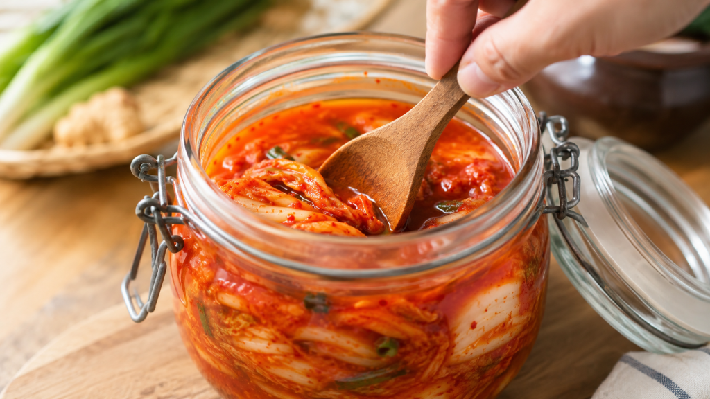 Một bàn tay đang dùng thìa gỗ nén chặt những miếng kim chi xuống dưới mặt nước sốt trong một hũ thủy tinh sạch sẽ. Hình ảnh này minh họa trực quan cho "mẹo" giữ kim chi ngon lâu trong tủ lạnh.