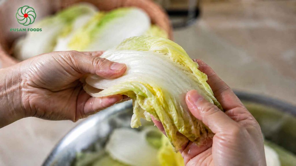 Một đôi tay đang cầm một bẹ cải thảo đã muối, bẻ cong lại để cho người đọc thấy cải đã dẻo, không bị gãy (đúng như mô tả trong bài). Đây là yếu tố "human touch" cực kỳ quan trọng để tăng uy tín cho bài hướng dẫn.